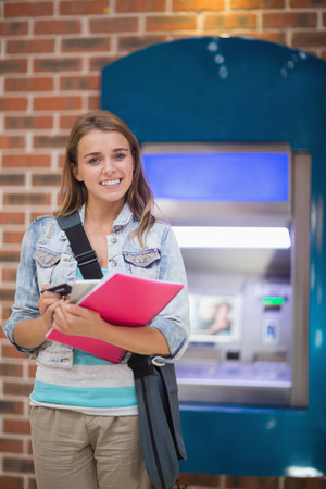 Pretty student standing smiling at camera at the atm in collegeの写真素材