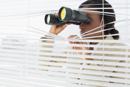 Serious young businesswoman peeking with binoculars through blinds in the officeの写真素材