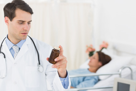Serious doctor holding a bottle of pills with patient in background at hospitalの写真素材