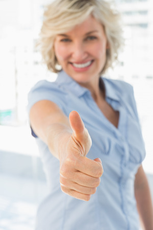 Portrait of an elegant businesswoman gesturing thumbs up in a bright officeの写真素材