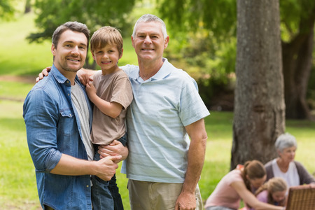 Portrait of grandfather, father and son with family in background at the parkの写真素材