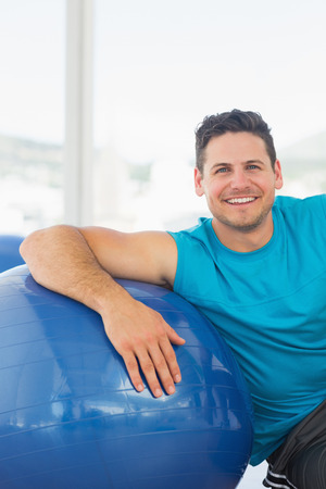 Portrait of a smiling young man sitting with fitness ball at a bright gymの写真素材