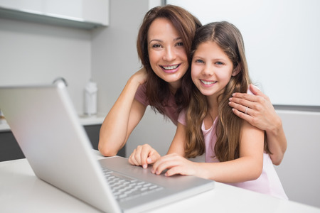 Portrait of a smiling mother with young daughter using laptop in the kitchen at home