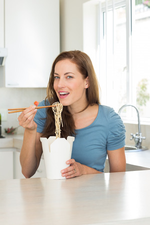 Portrait of a smiling young woman eating noodles in the kitchen at homeの写真素材