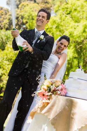 Happy young newlywed couple with groom opening the champagne bottle at parkの写真素材