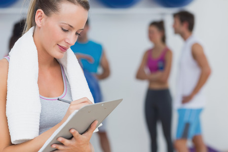 Female trainer writing on clipboard with fitness class in background at the gym