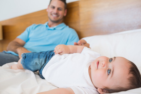 Smiling baby boy lying on bed with father in background の写真素材