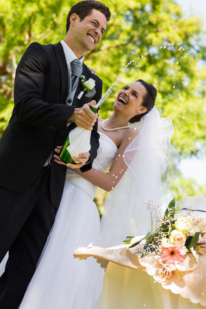 Happy young newlywed couple with groom opening the champagne bottle at parkの写真素材