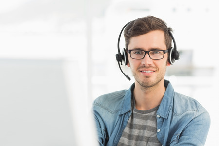 Portrait of a smiling casual young man with headset using computer in a bright office