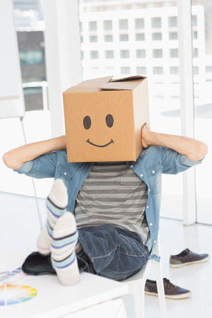 Casual young man with happy smiley box over face while sitting on chair at officeの写真素材