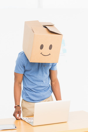 Casual young man with happy smiley box over face in front of laptop at officeの写真素材