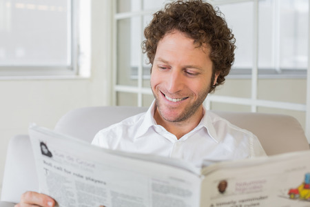 Closeup of a smiling young man reading newspaper on sofa in the houseの写真素材