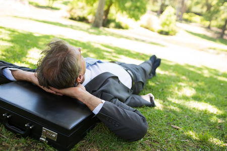 Businessman resting head on briefcase while lying in parkの写真素材