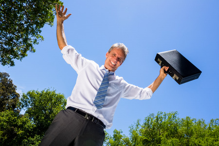 Low angle portrait of happy businessman with arms outstretched carrying briefcase against blue skyの写真素材