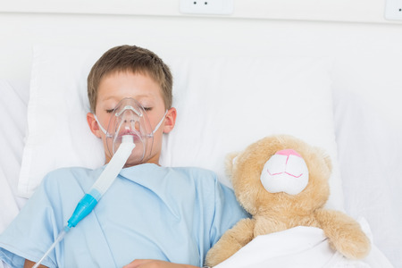 Sick boy wearing oxygen mask sleeping beside stuffed toy in hospital bedの写真素材