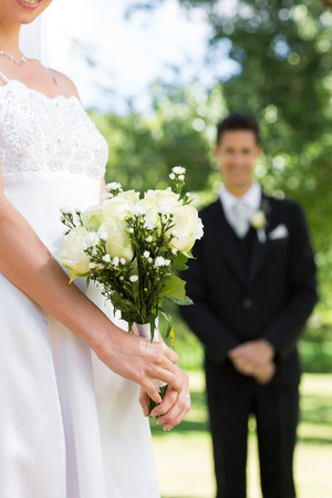 Midsection of bride holding bouquet with groom standing in background at gardenの写真素材