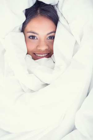 Pretty girl wrapped up in her duvet smiling at camera in her bedroom at homeの写真素材