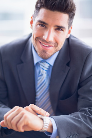Businessman sitting checking his watch smiling at camera in the officeの写真素材