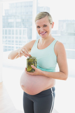 Smiling blonde pregnant woman eating from jar of pickles in a fitness studioの写真素材
