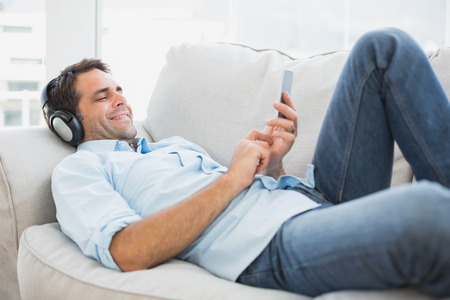 Happy handsome man lying on sofa using tablet and listening to music at home in the living roomの写真素材