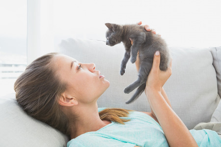 Cheerful woman lying on sofa holding a grey kitten at home in the living roomの写真素材