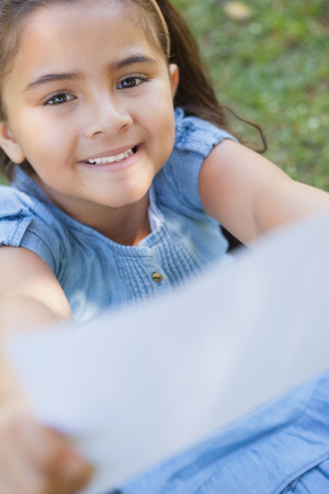 High angle portrait of a young smiling girl holding out a blurred paper at the parkの写真素材