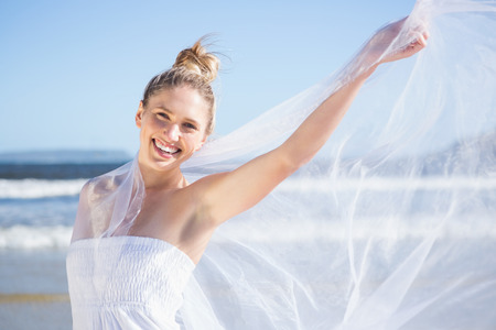 Pretty blonde in white dress holding up shawl on the beach on a bright dayの写真素材