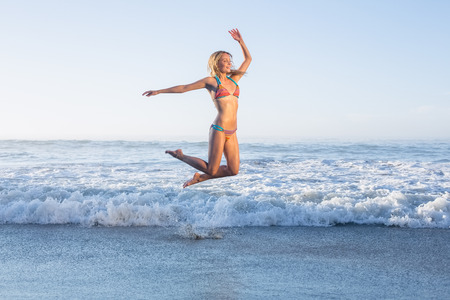 Happy blonde leaping on the beach in bikini on a sunny dayの写真素材