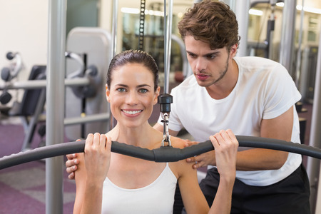 Fit brunette using weights machine for arms with trainer helping smiling at camera at the gym