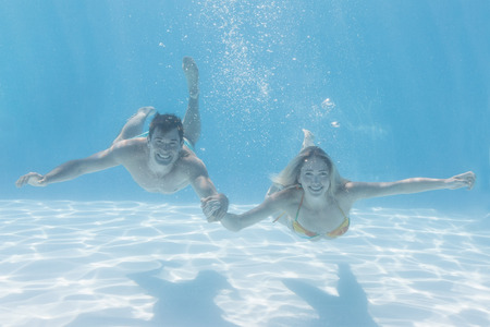 Cute couple smiling at camera underwater in the swimming pool on their holidaysの写真素材