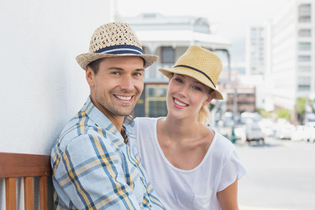 Young hip couple sitting on bench smiling at camera on a sunny day in the cityの写真素材