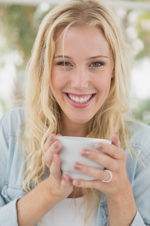 Pretty blonde sitting at table having coffee smiling at camera on the cafe terrace on sunny dayの写真素材