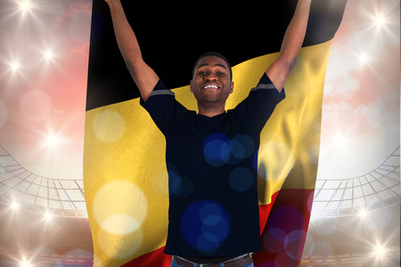 Excited football fan in black cheering holding belgium flag against large football stadium under cloudy blue skyの写真素材