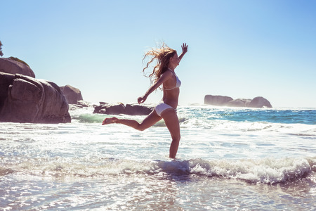 Beautiful smiling woman in white bikini skipping on the beach on a sunny dayの写真素材