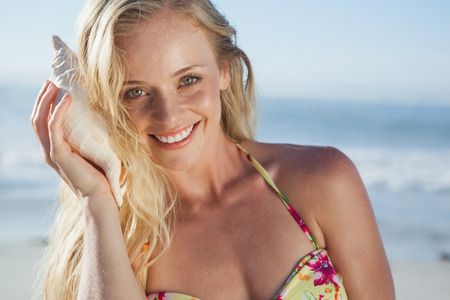 Pretty blonde in bikini listening to conch on the beach on a sunny dayの写真素材