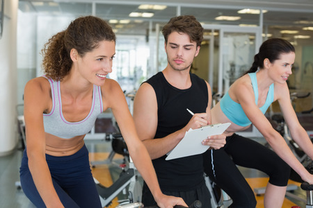 Fit women in a spin class with trainer taking notes at the gym