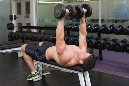 Shirtless bodybuilder lying on bench lifting heavy dumbbells at the gymの写真素材