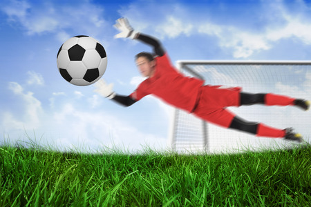 Fit goal keeper jumping up saving ball against field of grass under blue skyの写真素材
