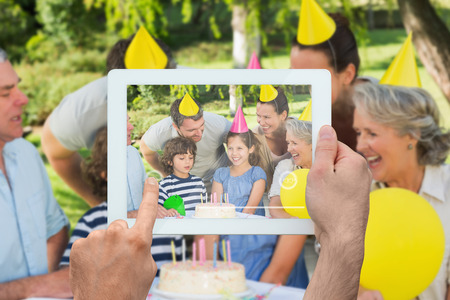 Hand holding tablet pc showing extended family wearing party hats at birthday celebration in park