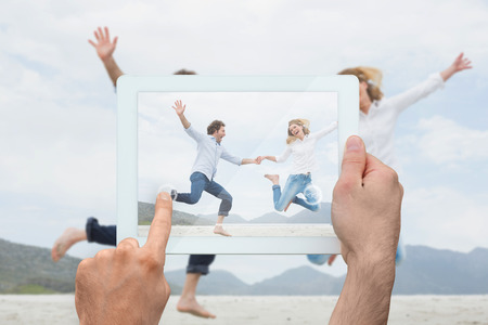 Hand holding tablet pc showing cheerful couple holding hands and jumping at beachの写真素材