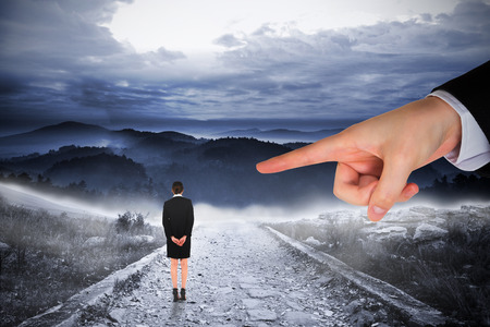 Giant hand pointing at businesswoman standing with hands behind back against stony path leading to large misty mountainsの写真素材