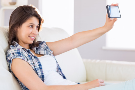 Pretty brunette taking a selfie on couch at home in the living roomの写真素材