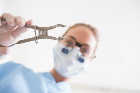 Dentist in surgical mask and dental loupes holding pliers over patient at the dental clinicの写真素材