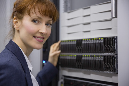 Pretty technician smiling at camera beside server tower in large data centerの写真素材