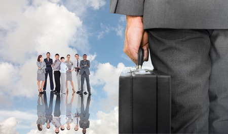 Businessman holding briefcase against blue sky with white clouds and business peopleの写真素材