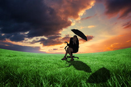 Young businesswoman holding umbrella against green field under orange skyの写真素材