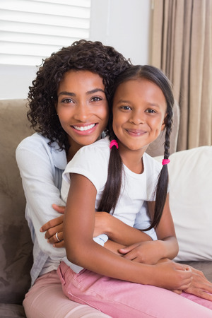 Pretty mother sitting on the couch with her daughter smiling at camera at home in the living roomの写真素材