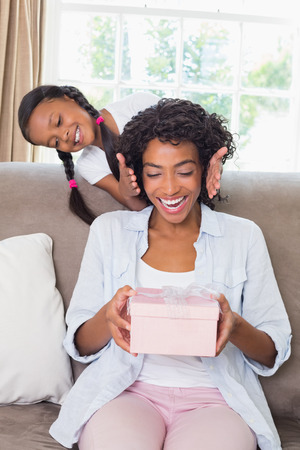 Pretty mother sitting holding gift with her daughter revealing at home in the living roomの写真素材