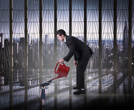 Businessman watering tiny businesswoman against room with large window looking on cityの写真素材