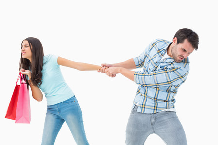 Attractive young man pulling his shopaholic girlfriend on white backgroundの写真素材
